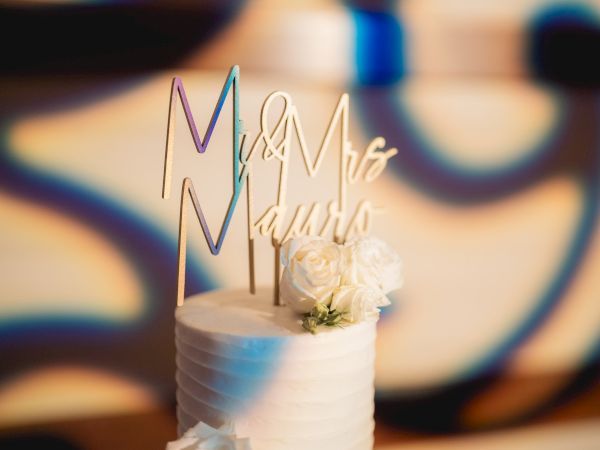 A white wedding cake with "Mr & Mrs" topper and white flowers against a blurred vibrant background.