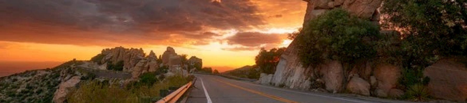 A scenic road with guardrails curves through rocky terrain, leading towards a vibrant sunset with dramatic cloud formations in the sky.