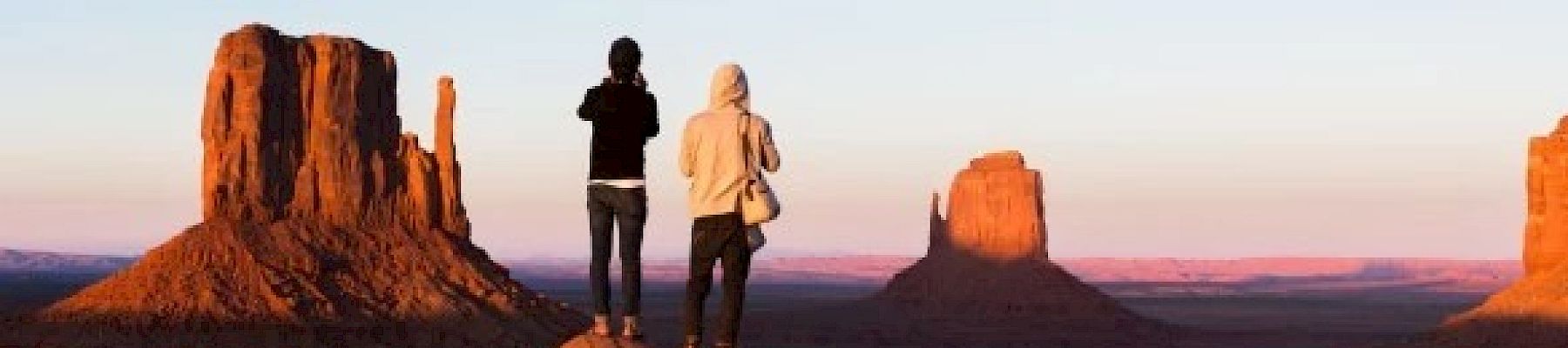 Two people stand on a rocky outcrop, overlooking iconic buttes at sunset in a desert landscape.