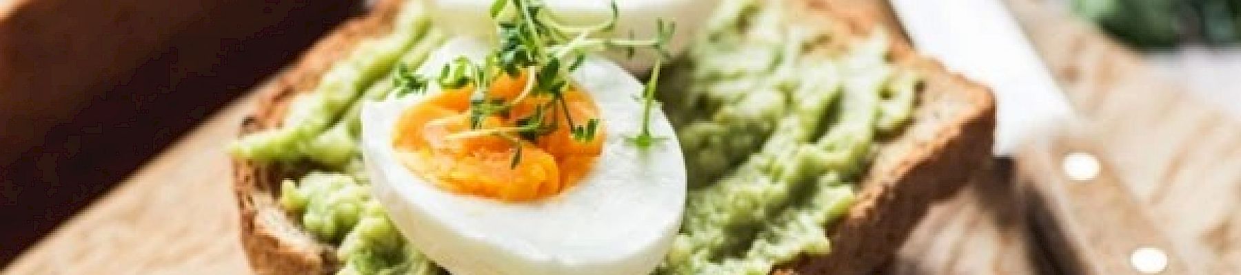 The image shows avocado toast topped with sliced boiled eggs and garnished with microgreens, placed on a wooden surface next to a knife.