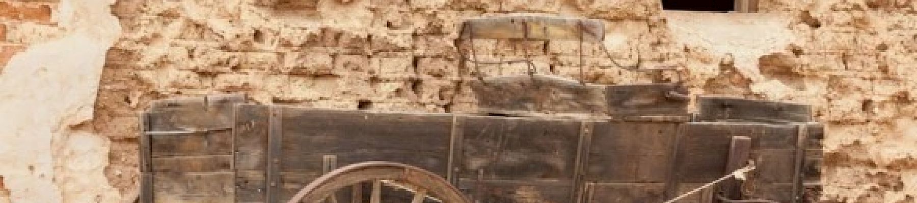 A wooden horse-drawn wagon with large spoked wheels stands against a deteriorating stone wall with a small window and no glass.