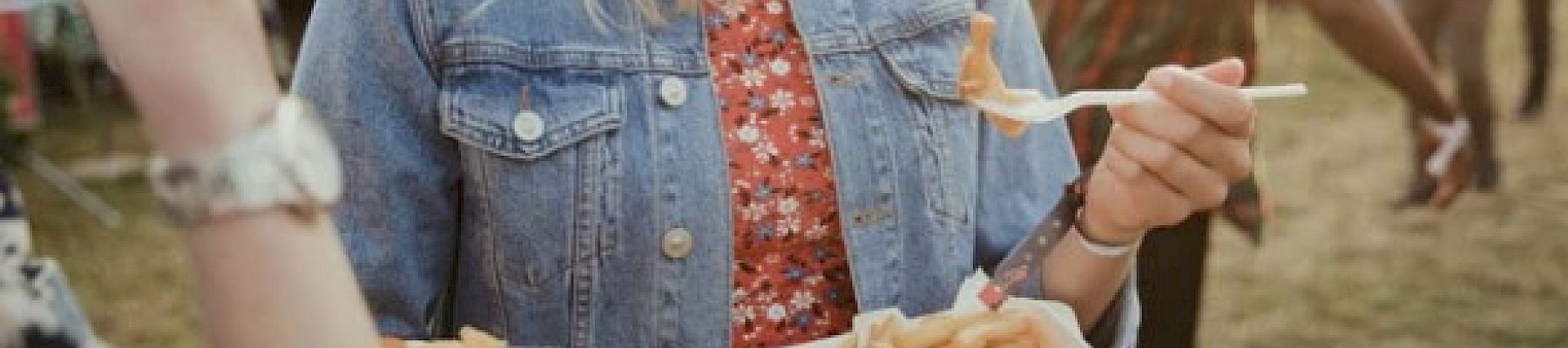 A person is smiling while holding a tray of fries at an outdoor event, wearing a denim jacket and a floral dress.