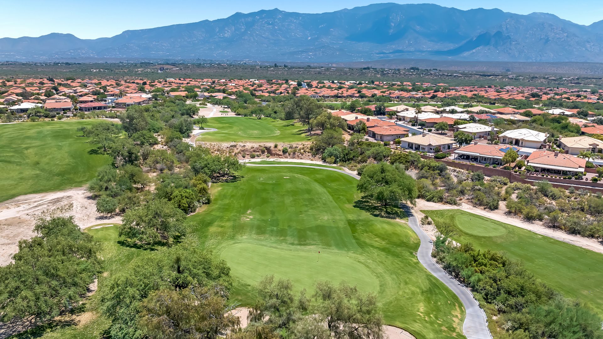 This image shows a golf course with green fairways, a residential area with numerous houses, and mountains in the background under a clear sky.