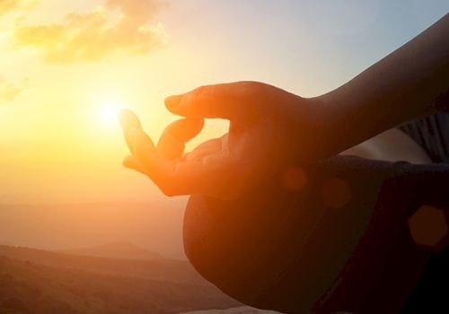 A person is meditating outdoors at sunset, with fingers forming a mudra gesture, against a scenic background with the sun setting behind them.