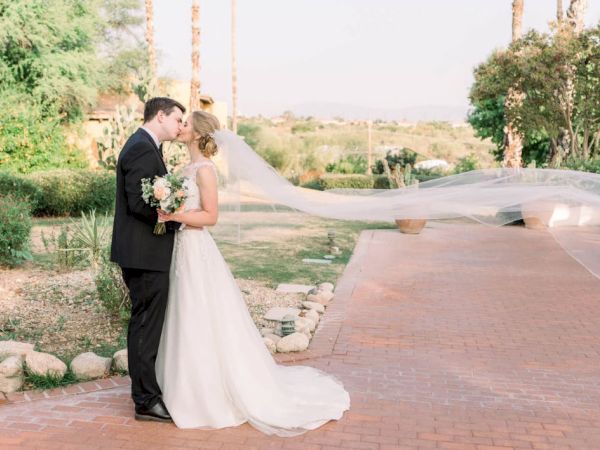 A bride and groom are kissing outdoors, with the bride's veil flowing in the breeze, surrounded by greenery and a brick path.
