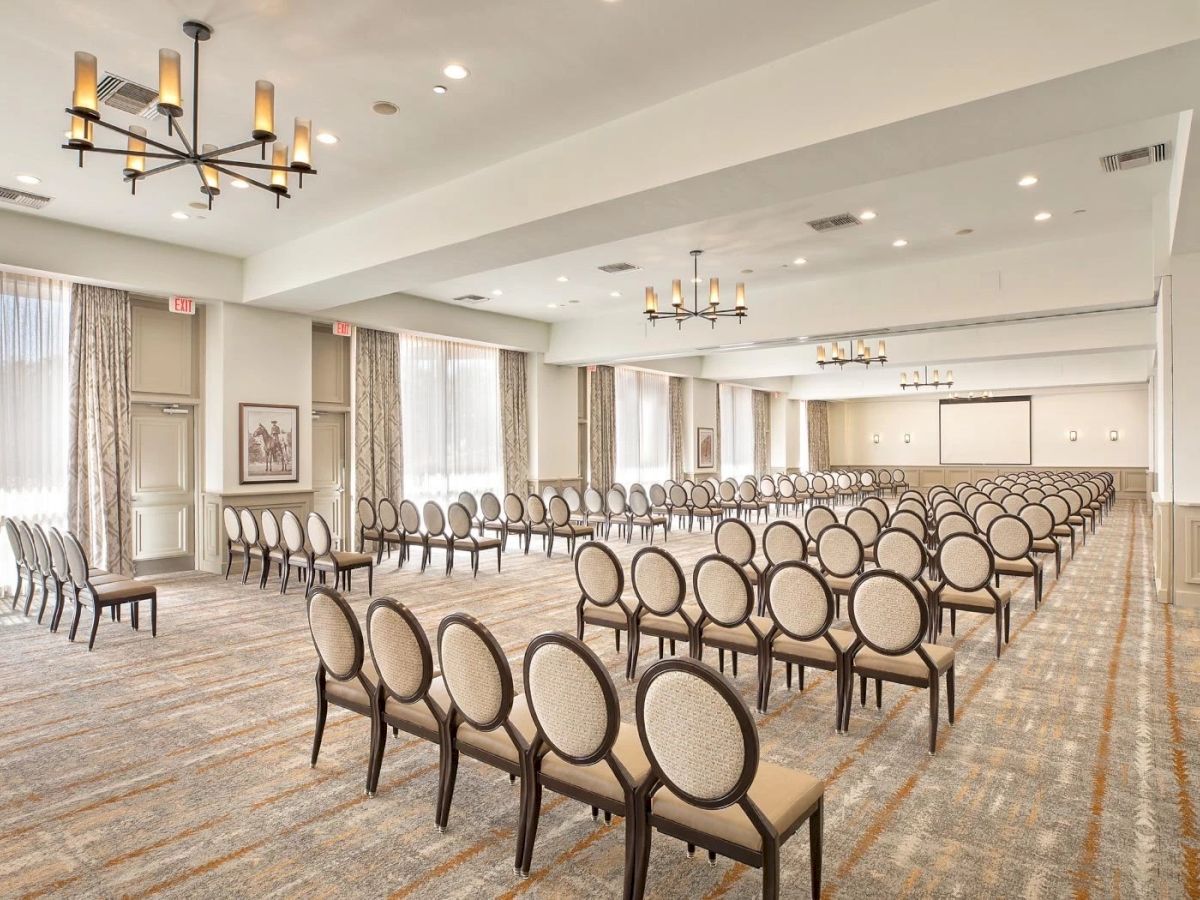 The image shows a large, well-lit conference room set up with rows of chairs facing a white screen, suitable for a meeting or presentation.