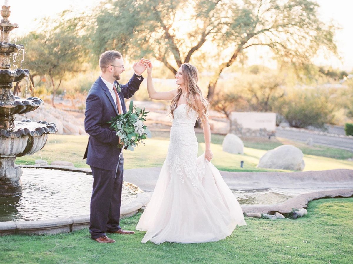 A couple, dressed formally, dances near a fountain in a park with trees and rocks in the background. The scene appears to be during sunset.