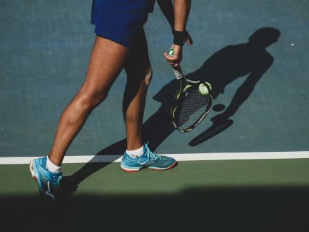 A tennis player in blue shorts and sneakers is preparing to serve on a court, with their shadow and the ball visible on the ground.