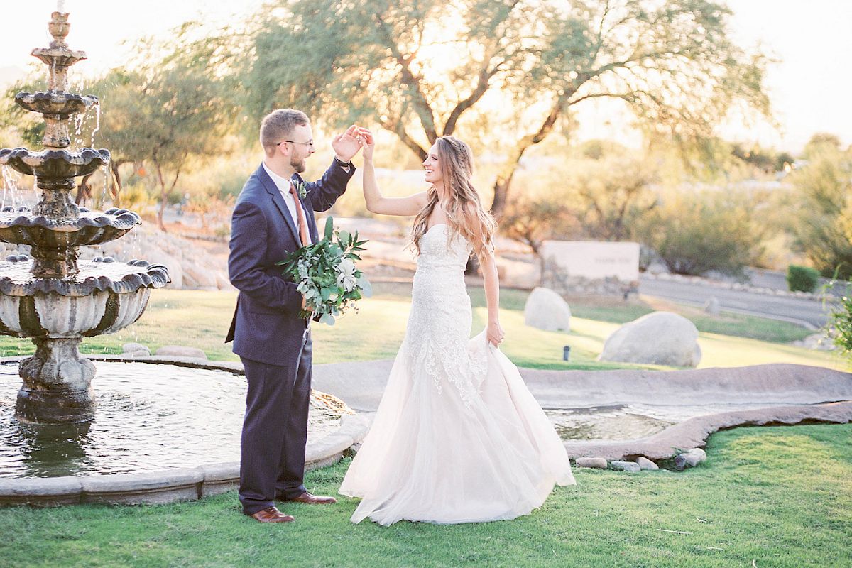 A couple in wedding attire dances near a fountain in a scenic outdoor setting with greenery and trees in the background.