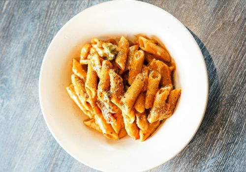 A bowl of penne pasta topped with a reddish sauce and black pepper, placed on a wooden table.