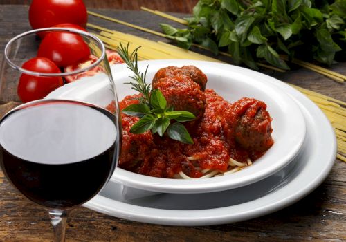 A plate of spaghetti with meatballs in tomato sauce, garnished with herbs, near a glass of red wine and fresh ingredients.