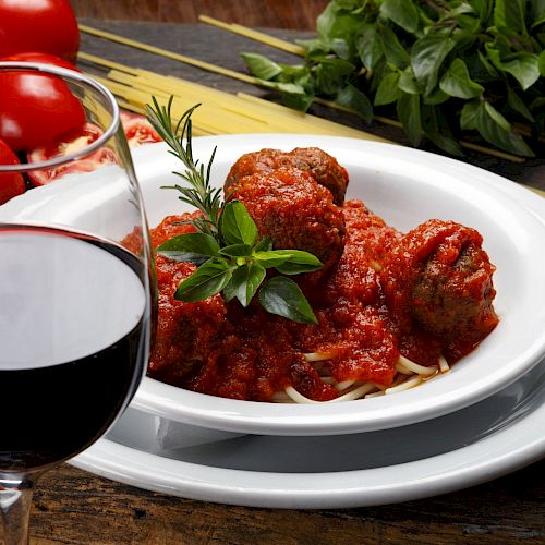 A plate of spaghetti with meatballs in tomato sauce, garnished with herbs, near a glass of red wine and fresh ingredients.