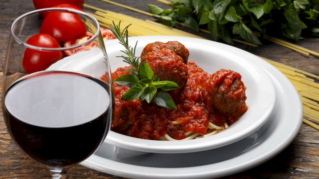 A plate of spaghetti with meatballs in tomato sauce, garnished with herbs, near a glass of red wine and fresh ingredients.