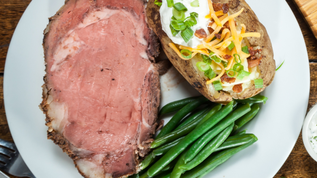 The image shows a plate with a serving of prime rib, a baked potato topped with sour cream, cheese, bacon, and green onions, alongside green beans.