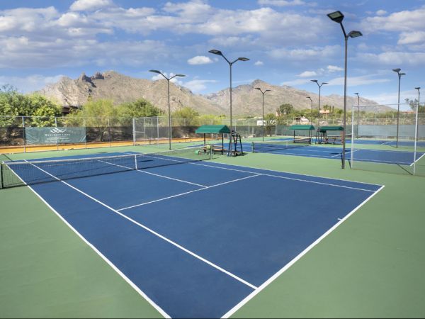 The image shows multiple outdoor tennis courts with blue and green surfaces, surrounded by desert vegetation and mountains in the background.