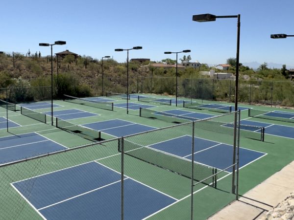 The image shows a fenced outdoor area with multiple blue and green pickleball courts. The area is well-lit with numerous tall lights.