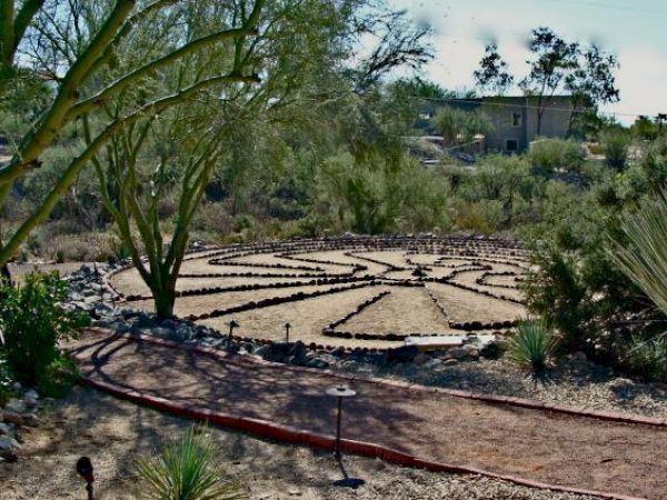 A garden scene with a labyrinth made of stones, surrounded by trees and plants, with a house in the background.