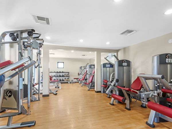 The image shows a well-equipped gym with various workout machines, including weight machines and leg press equipment, against a light-colored wall.
