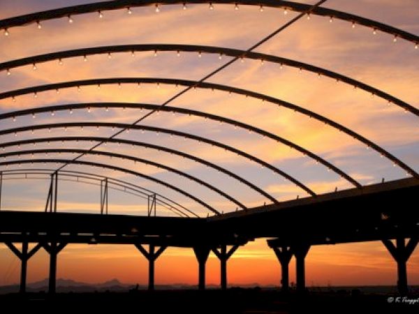 The image shows a silhouetted structure with curved beams against a backdrop of a colorful sunset, with lights hanging from the beams.