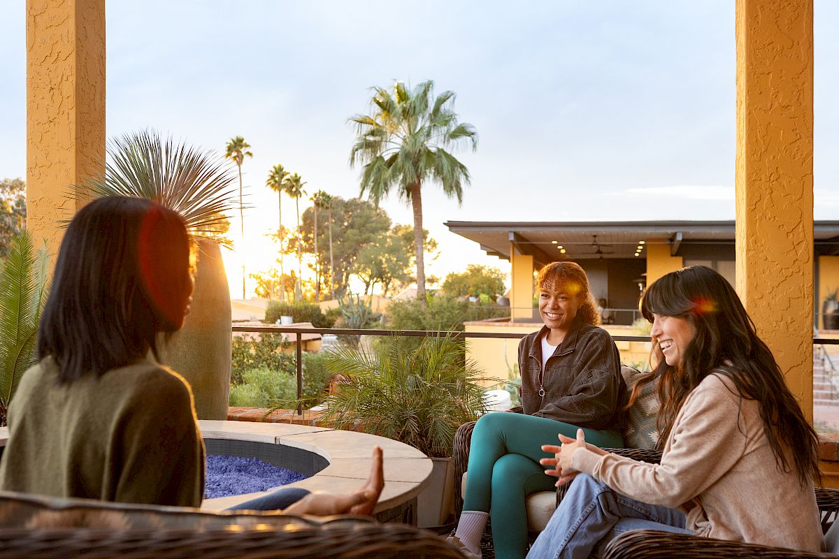 Friends chat on a sunny patio at sunset, surrounded by palm trees.