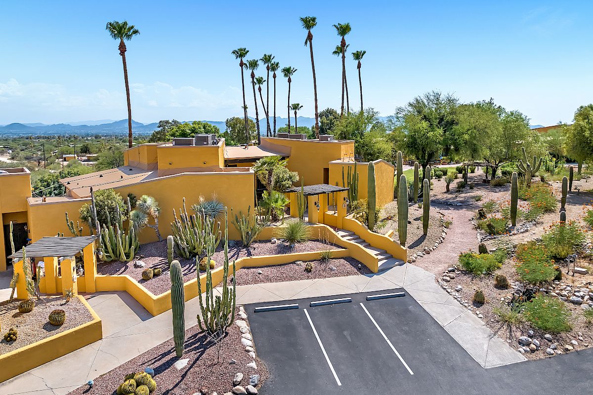 The image depicts a yellow building complex surrounded by desert landscaping, featuring cacti and palm trees, with a parking area in the foreground.