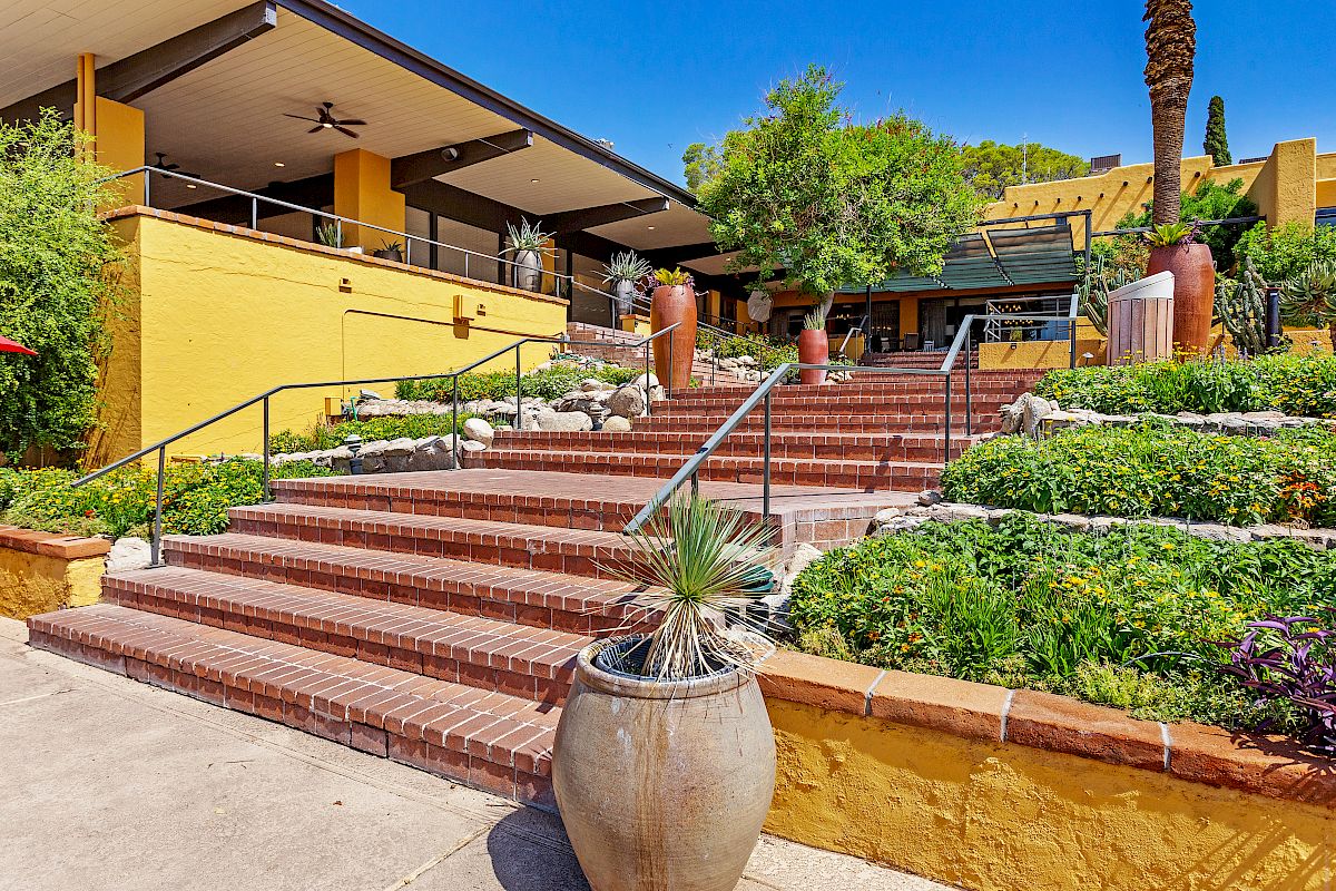 The image shows an outdoor stairway leading to a building entrance, surrounded by lush greenery, large potted plants, and modern architectural elements.