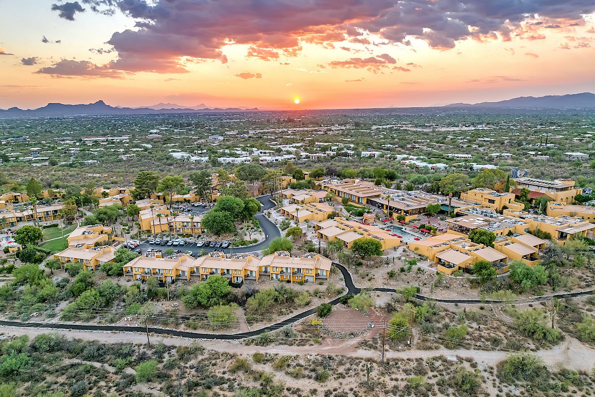An aerial view of a residential area with multiple buildings, surrounded by greenery and a desert landscape, with a sunset in the background.