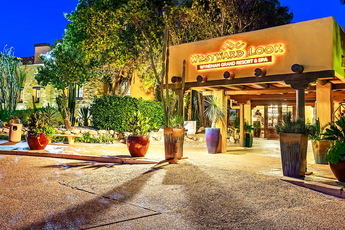 A well-lit building entryway with a sign that reads "Westward Look Wyndham Grand Resort & Spa" surrounded by greenery and potted plants.
