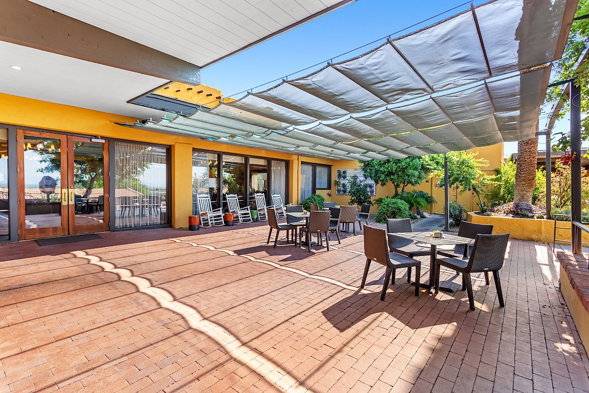 An outdoor patio area with several tables and chairs, covered by a shade structure. The building has large windows and plants nearby.