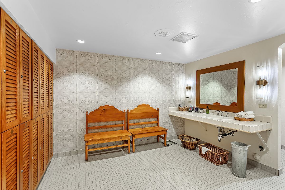 The image shows a well-lit bathroom with wooden benches, a large countertop with a sink, a mirror, and wicker baskets underneath.