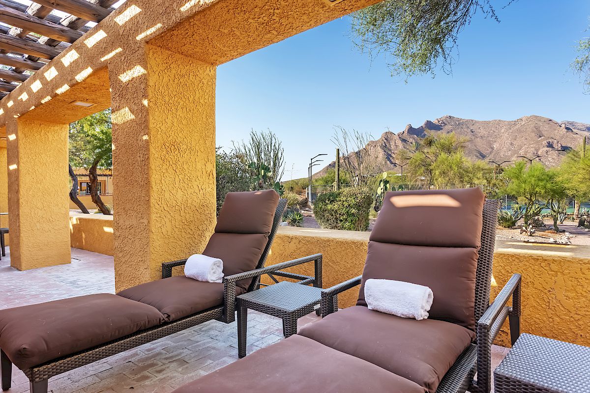 The image shows two lounge chairs with towels rolled on top, under a pergola with a scenic mountain view and desert vegetation.