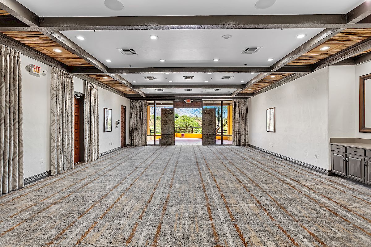 An empty, modern conference room with carpeted floor, exposed ceiling beams, and large glass doors leading to an outdoor view.