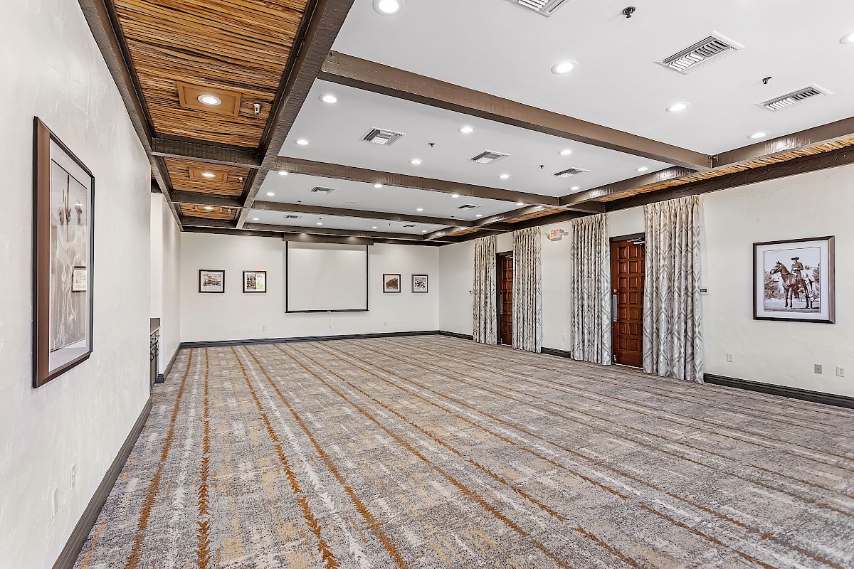 An empty conference room with framed pictures on the walls, carpeted floor, wood-beamed ceiling, and a projection screen at the front.