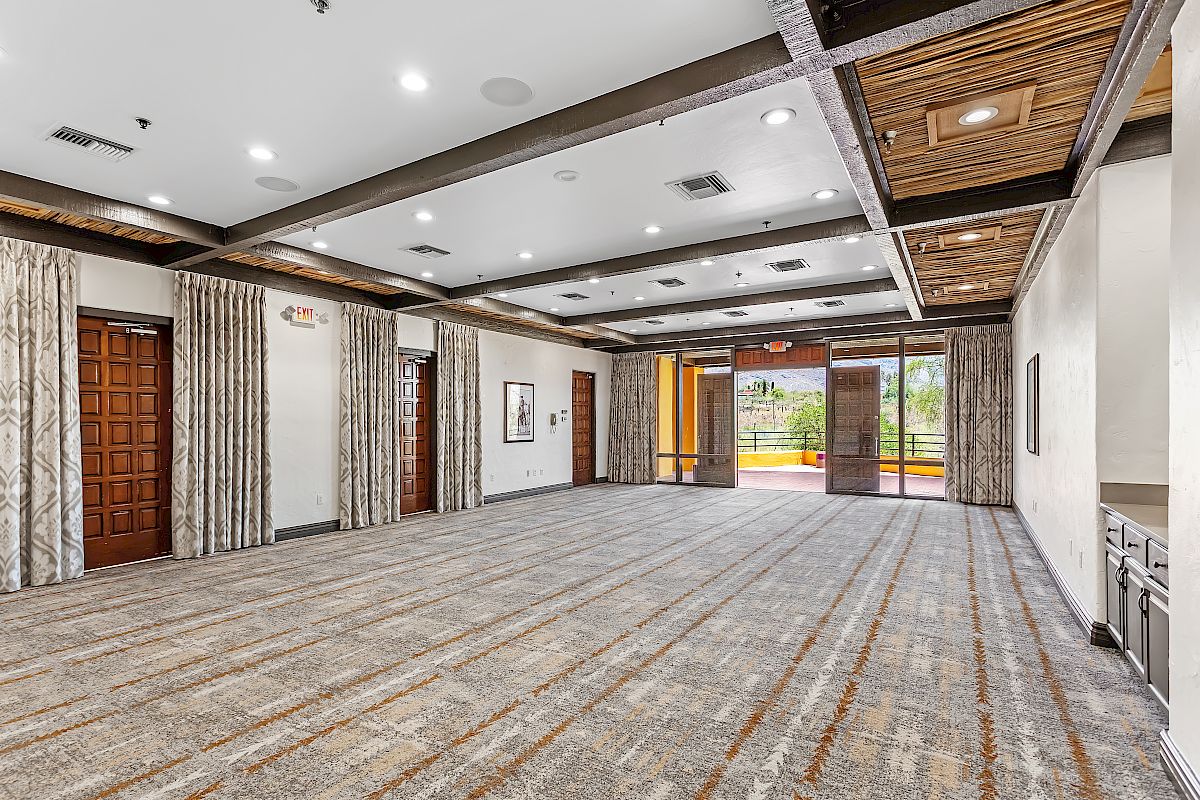 An empty, modern conference room with large windows, carpeted flooring, wooden ceiling beams, and double doors, featuring ample natural light.