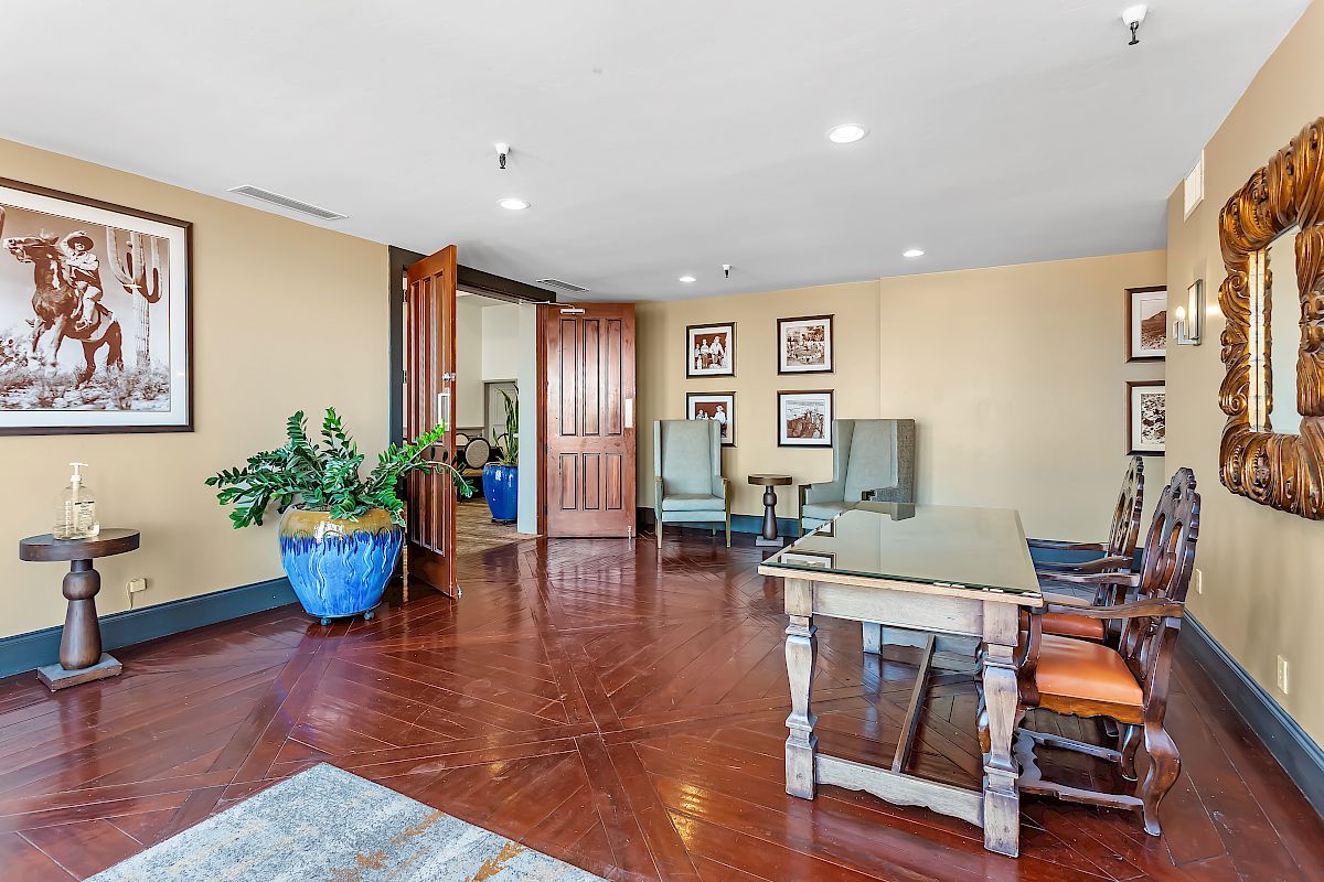 An elegantly decorated office with a wooden desk, chairs, wall art, a large plant, and a mirror on the wall, featuring hardwood floors and ample lighting.
