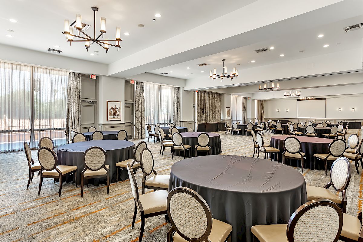 The image shows an empty banquet hall with several round tables covered in dark tablecloths and surrounded by chairs, with chandeliers hanging from the ceiling.