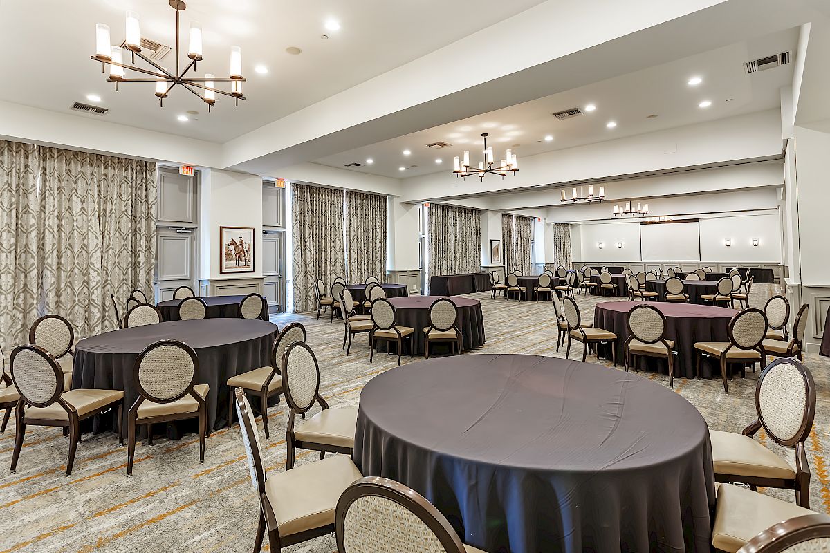 This image shows a spacious, well-lit event room with round tables covered with black tablecloths and chairs arranged around them, ready for an event.