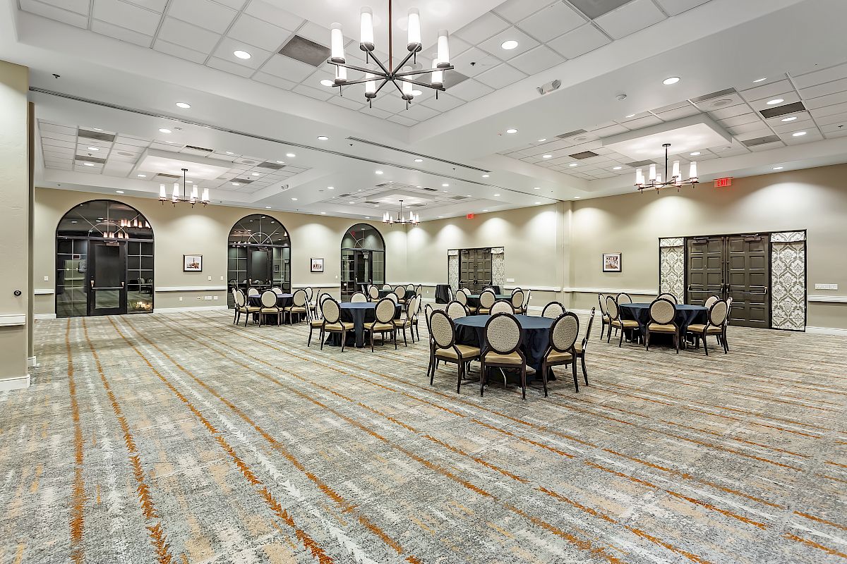 This image shows a large, empty banquet hall with round tables and chairs set up sparsely, chandeliers, and arched windows and doors.