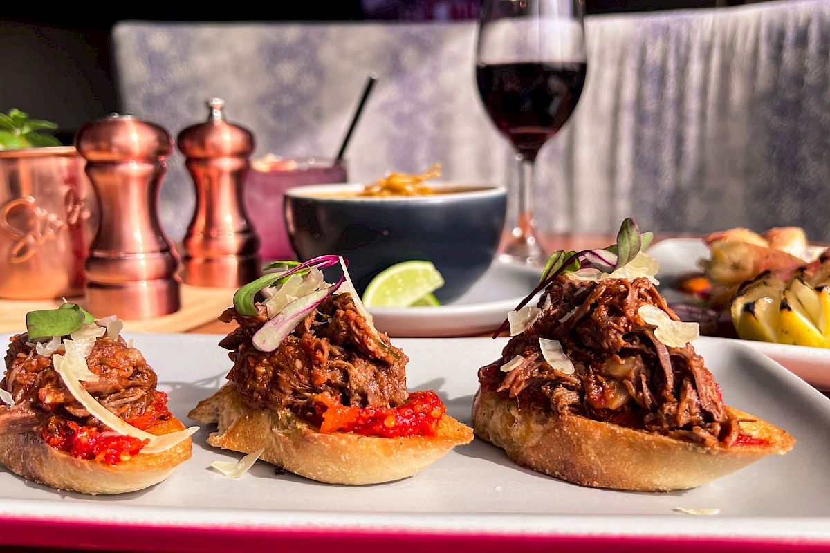 This image shows gourmet appetizers on toast with meat and garnish, accompanied by drinks, including a glass of red wine, and various other dishes.