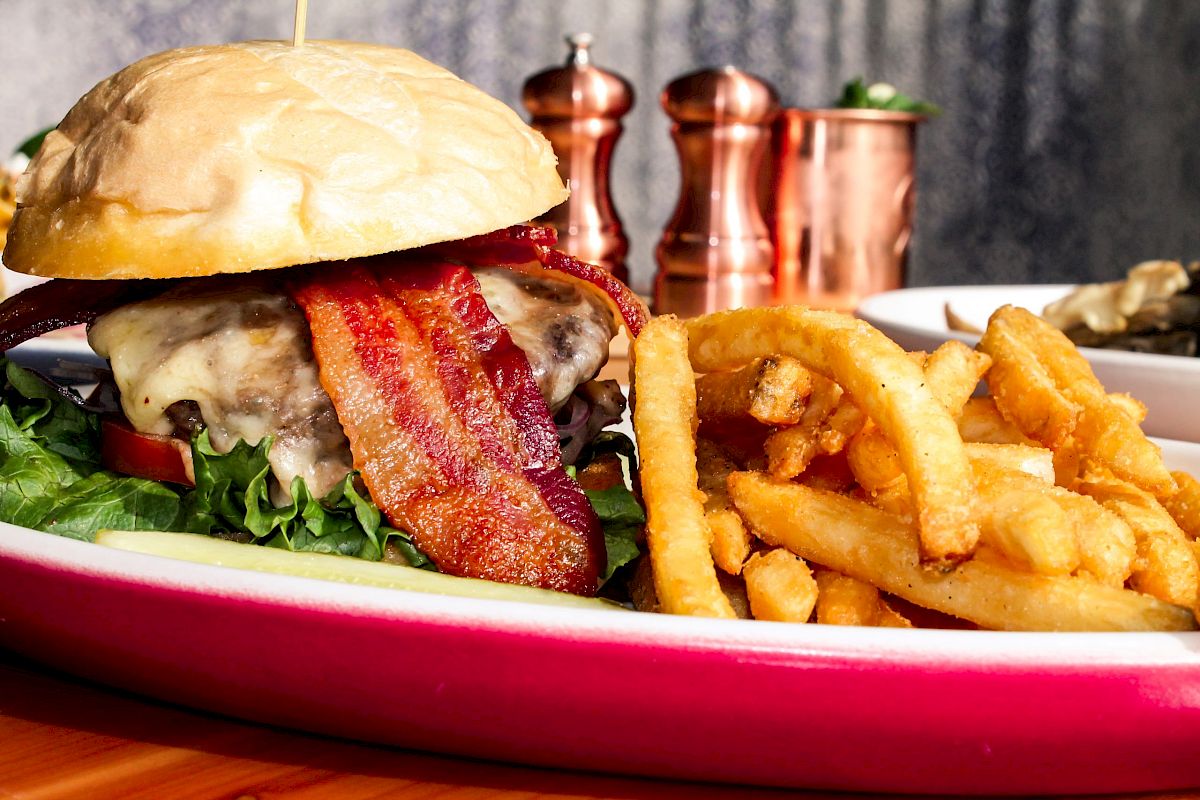 A close-up of a bacon cheeseburger with fries on a red-rimmed plate, with pepper shakers and a container in the background.