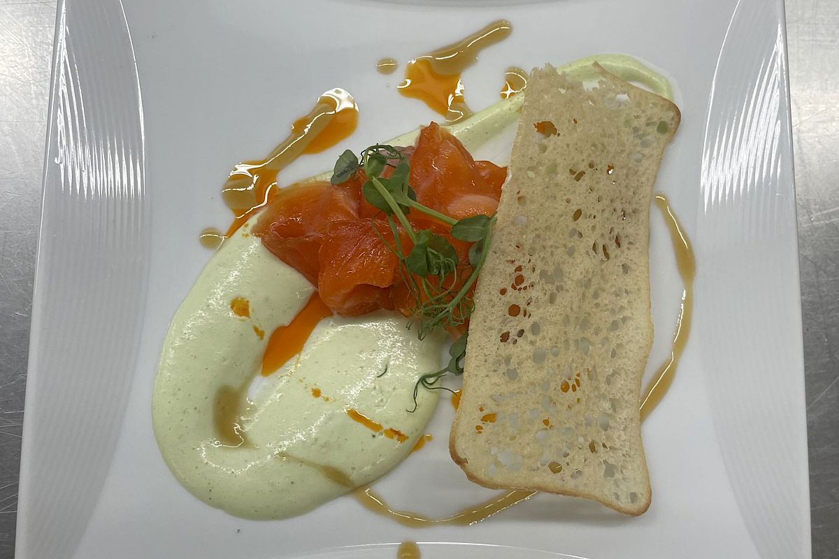 A plated dish featuring a slice of bread, creamy sauce, smoked salmon, herbs, and a drizzle of sauce on a square white plate.