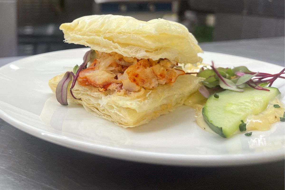 A plated dish featuring a puff pastry filled with lobster, accompanied by thin cucumber slices, red onions, and a garnish on the side.