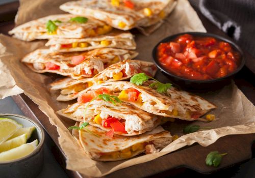 Quesadillas with cheese, corn, and peppers are on a tray, served with a side of salsa and lime wedges.