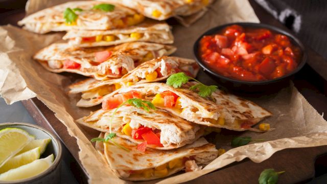 Quesadillas with cheese, corn, and peppers are on a tray, served with a side of salsa and lime wedges.