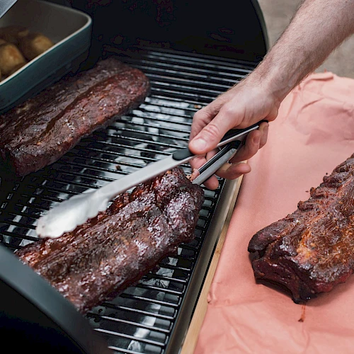 A person is grilling pieces of meat on a barbecue, using tongs to handle the food, with potatoes in a tray beside the meat.