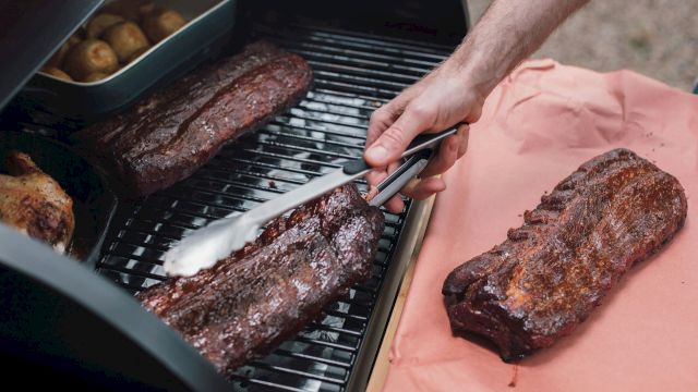 A person is grilling pieces of meat on a barbecue, using tongs to handle the food, with potatoes in a tray beside the meat.