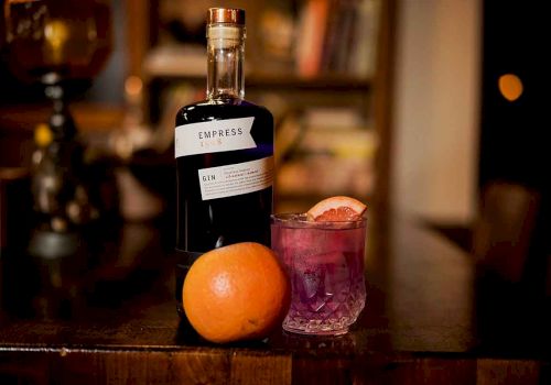 The image shows a bottle of Empress 1908 gin, a cocktail garnished with citrus, and an orange on a wooden table in a cozy setting.