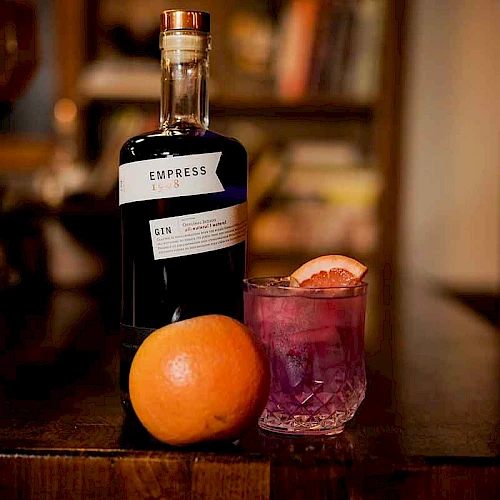 The image shows a bottle of Empress 1908 gin, a cocktail garnished with citrus, and an orange on a wooden table in a cozy setting.