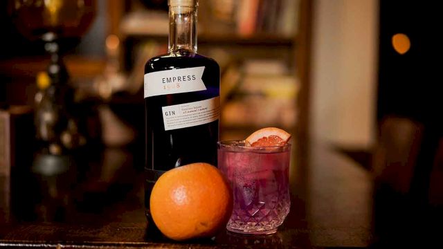 The image shows a bottle of Empress 1908 gin, a cocktail garnished with citrus, and an orange on a wooden table in a cozy setting.