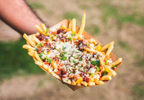 A hand holds a tray of loaded fries topped with cheese, bacon bits, and green onions over a grassy background.